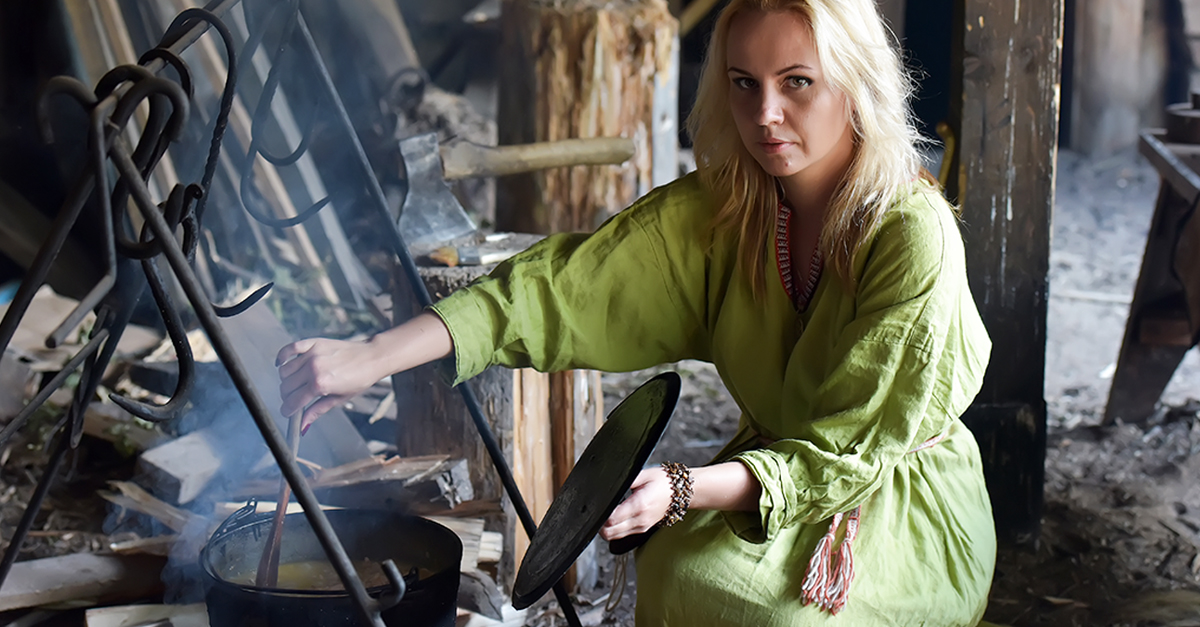 Blonde girl in vintage viking clothing prepares food in a cauldron on a fire